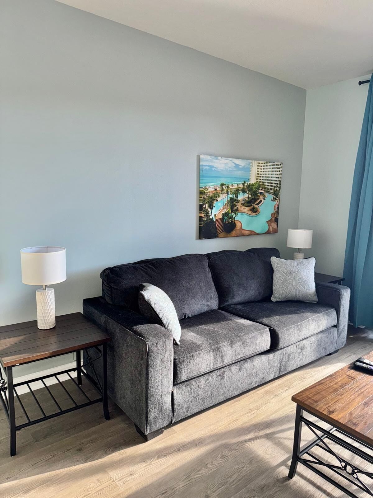 A living room with a grey sofa, two side tables with lamps, a wooden coffee table, and a painting of a resort on the wall.