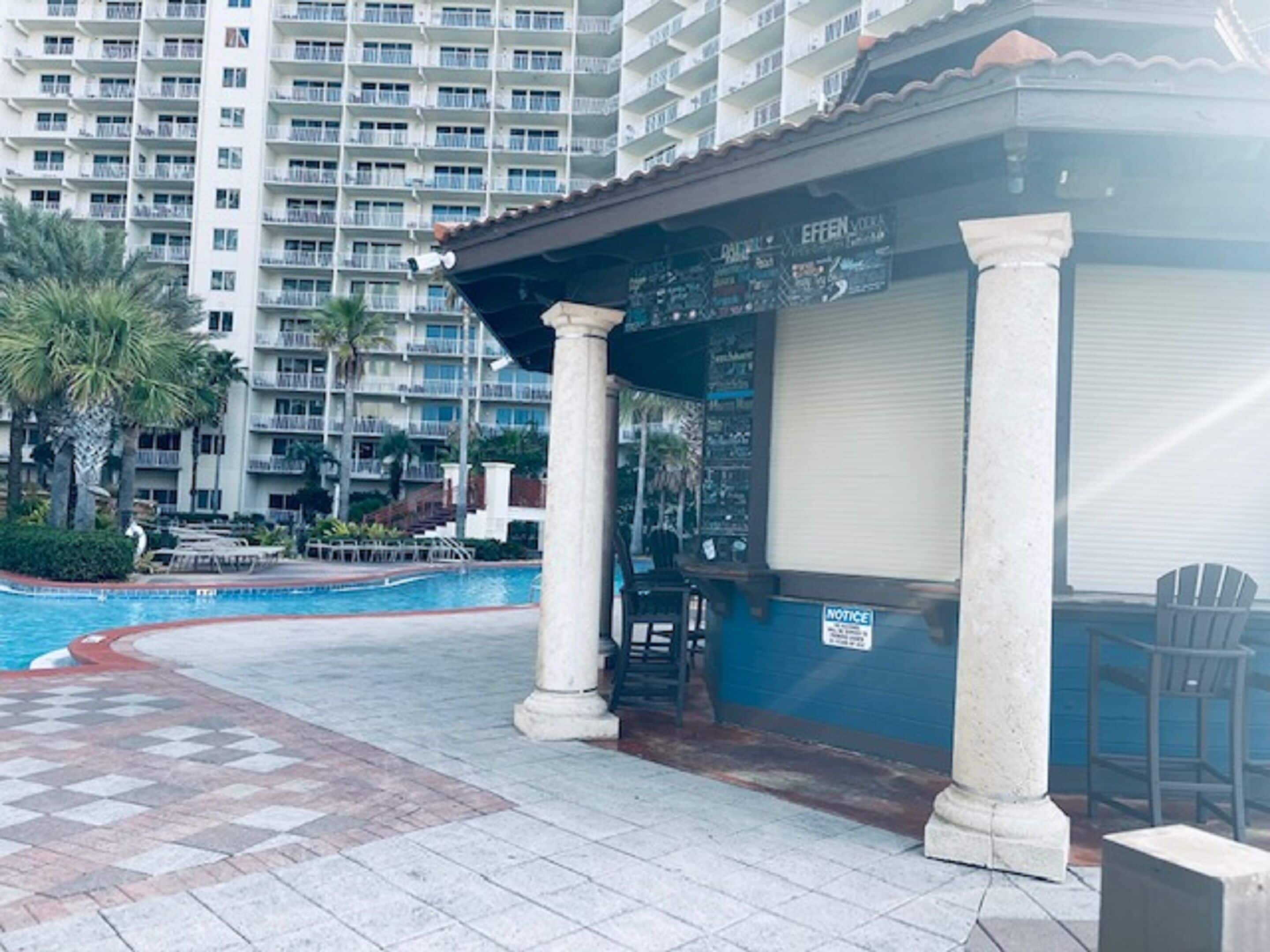 A pool area with a covered seating area, palm trees, and a multi-story building in the background.