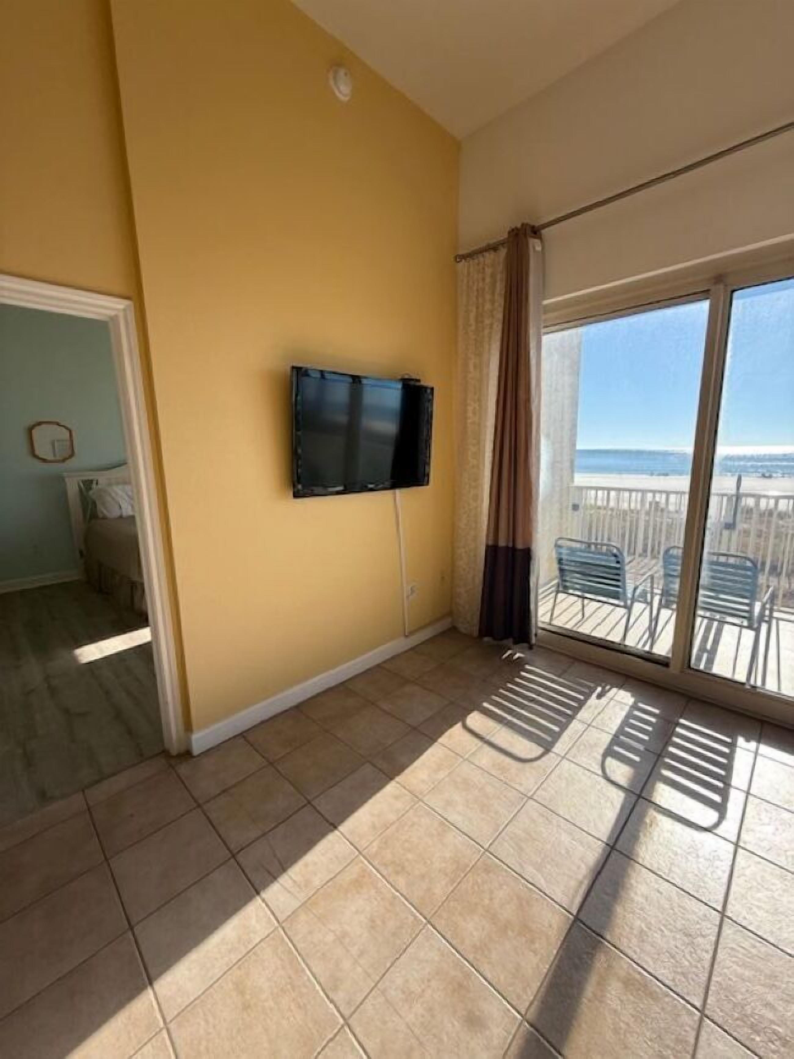 A room with a large window, a flat-screen TV mounted on the wall, and a view of the beach.
