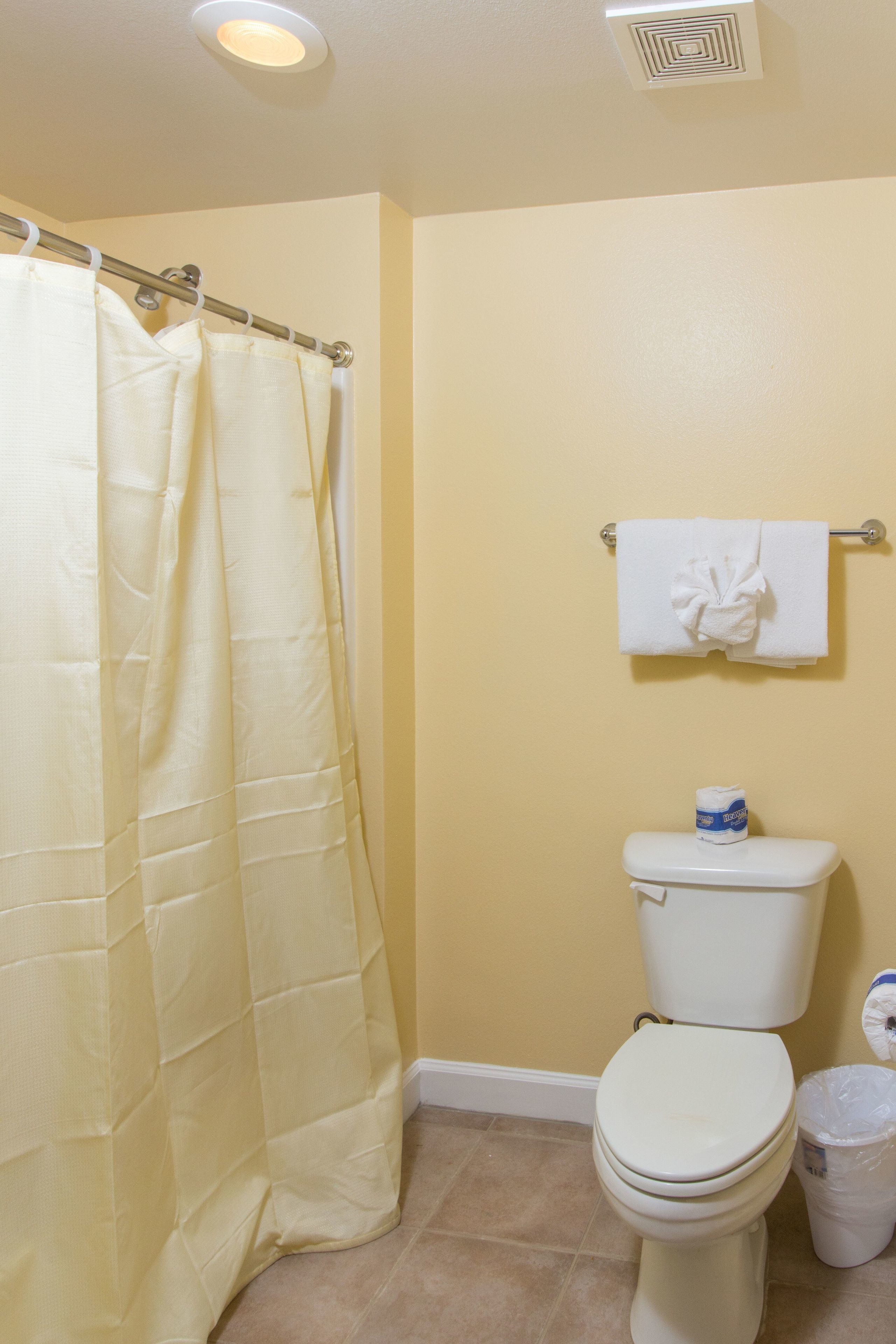 Un baño con una pared amarilla, un inodoro blanco, una toalla blanca colgada en un perchero y una cortina de ducha.