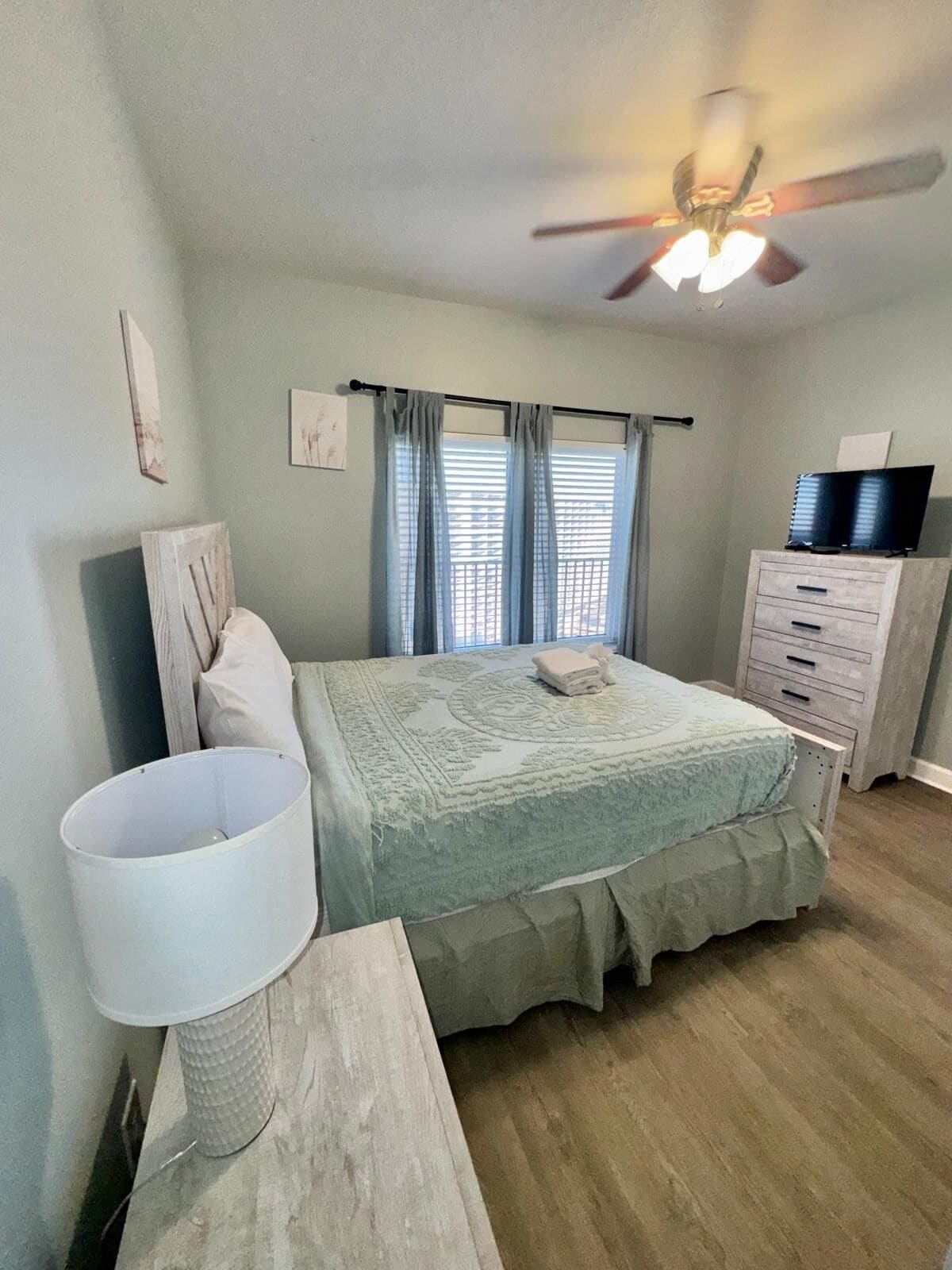 A bedroom with a bed, a dresser, a ceiling fan, and a window with curtains.