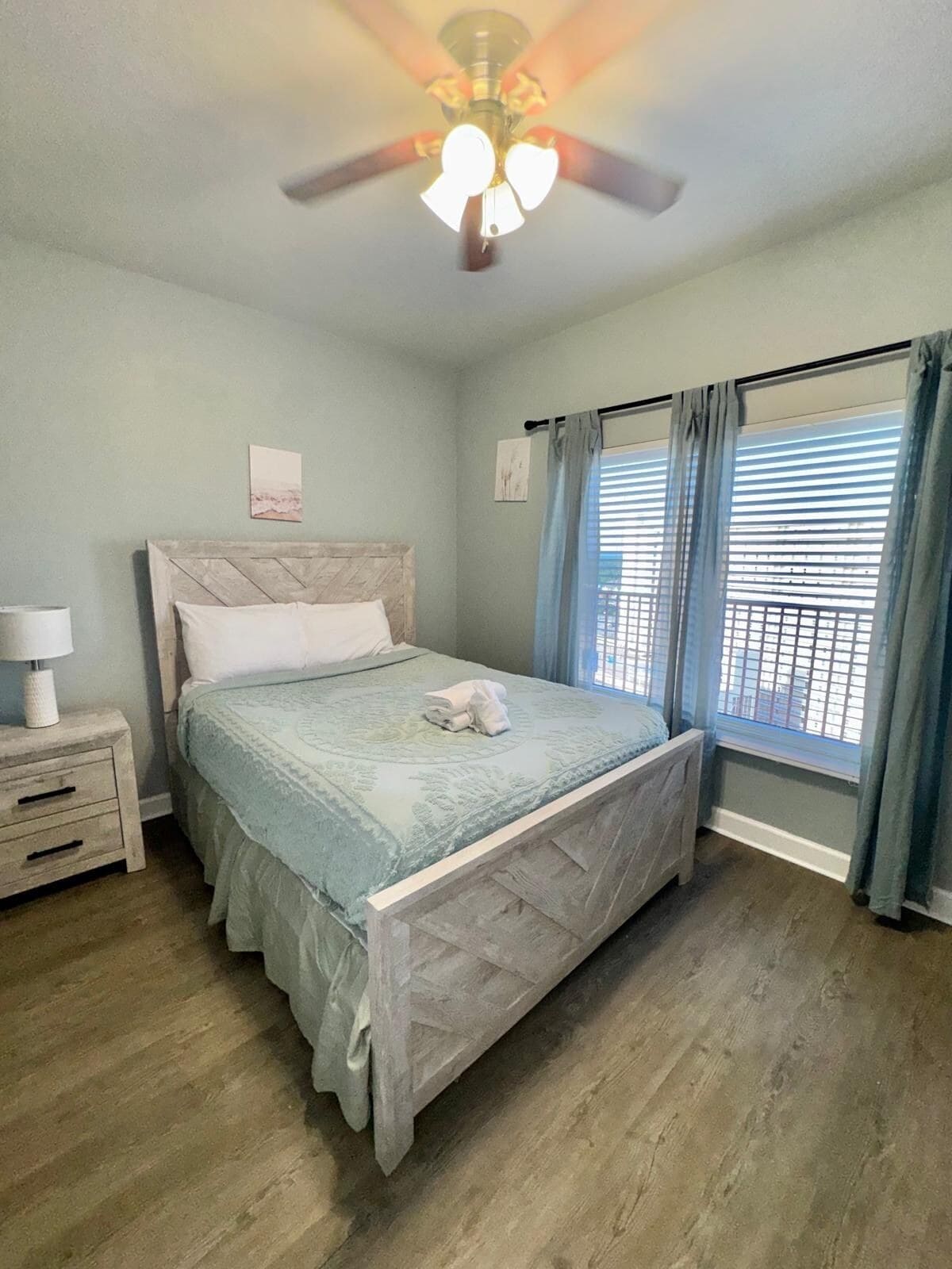 Une chambre à coucher comprenant un lit, une table de chevet, un ventilateur de plafond et une fenêtre avec des rideaux.