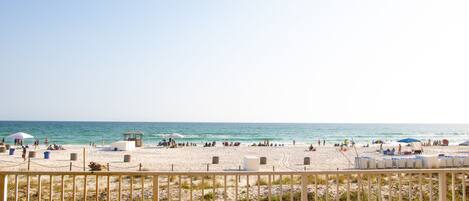 A beach with a wooden fence in the foreground, a lifeguard tower, and umbrellas in the background.