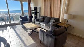 A spacious living room with a large window offering a view of the ocean, a sofa set, a coffee table, and a side table with a lamp.
