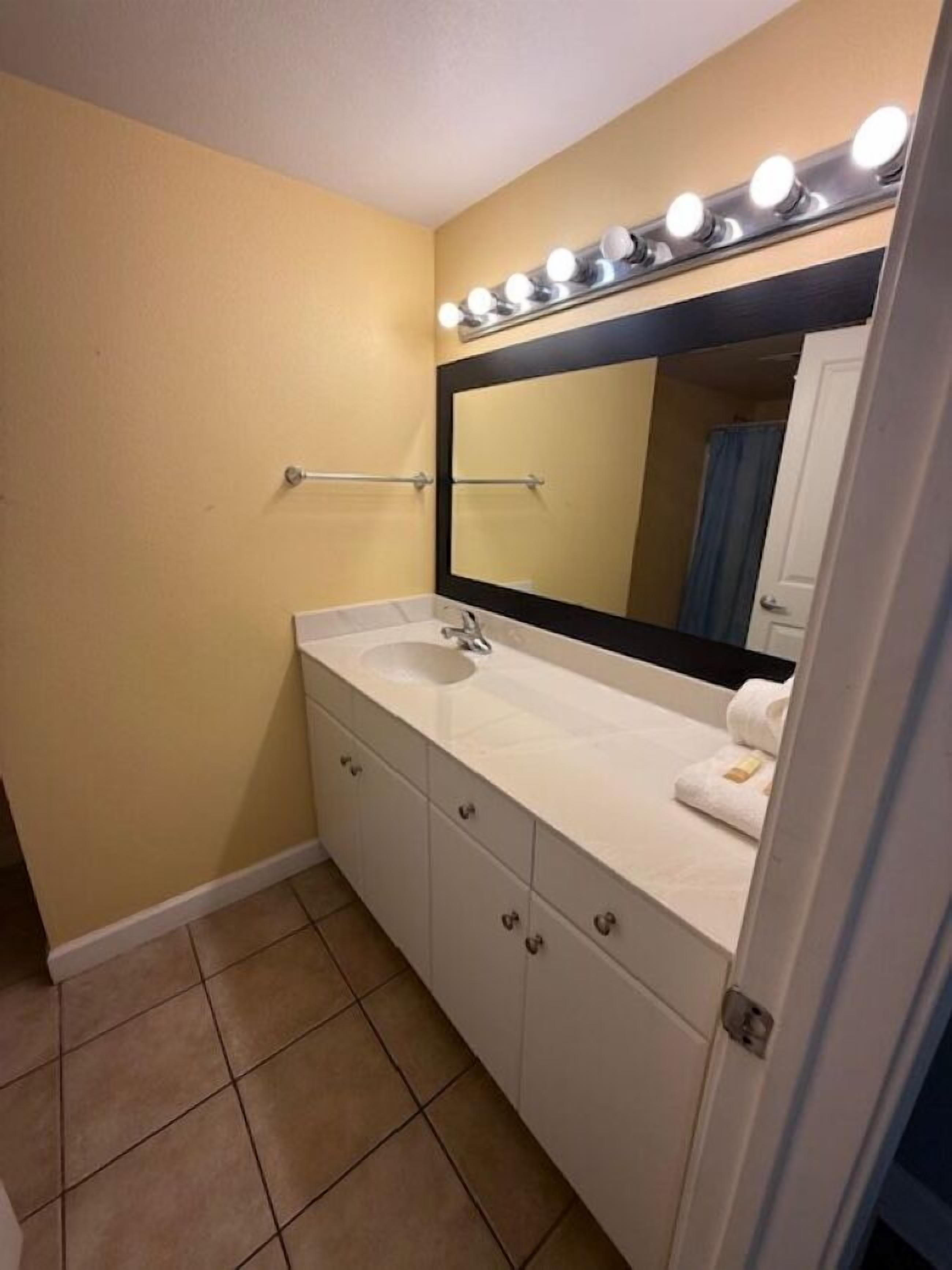 A bathroom with a white vanity, a large mirror with lights, and a towel rack.
