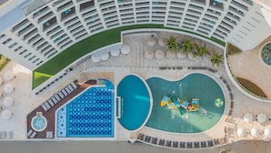 Vista aérea de un hotel con una piscina central grande, varias piscinas más pequeñas y una terraza en la azotea con asientos al aire libre.