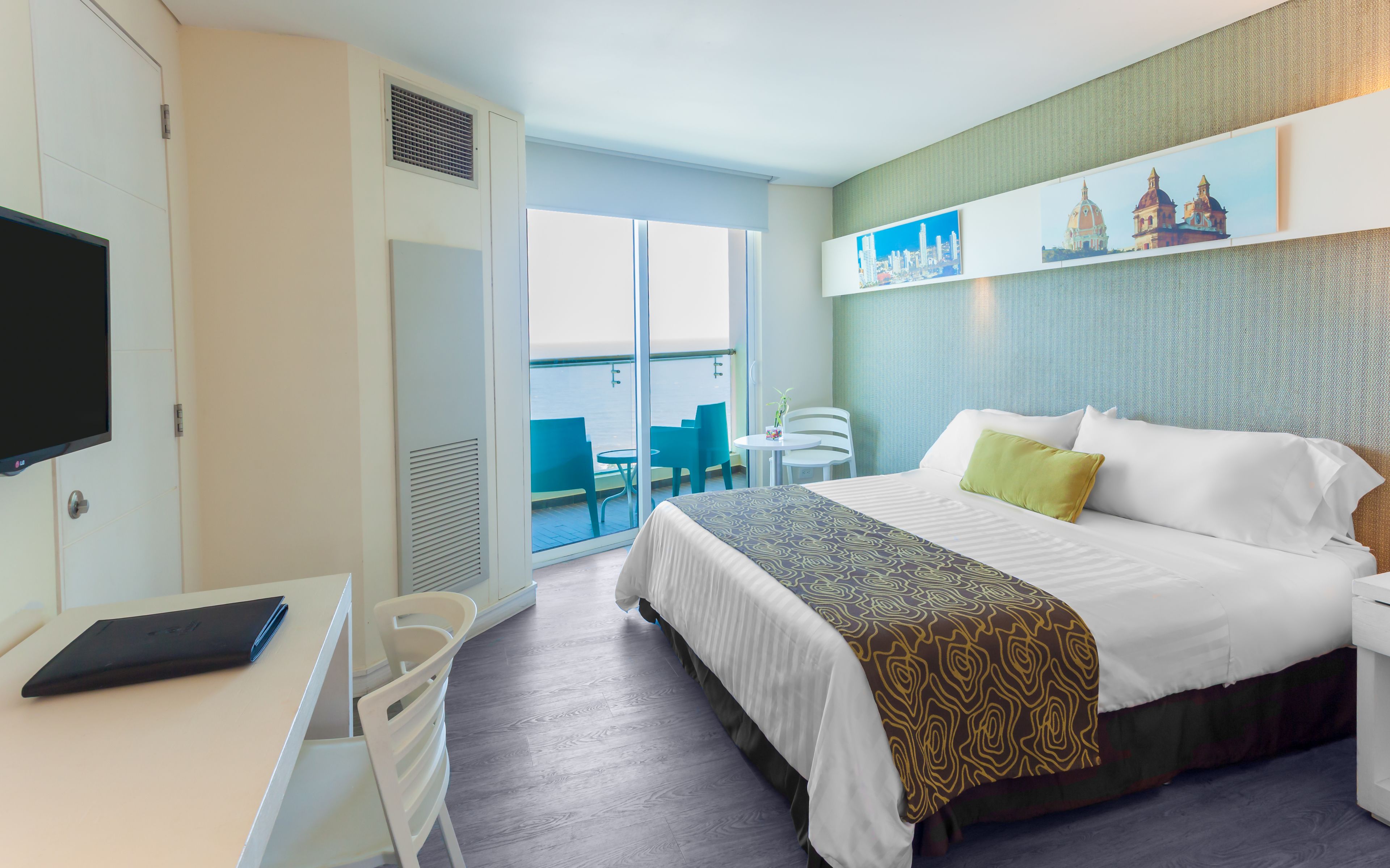 Habitación de hotel con cama, escritorio con computadora portátil, televisión, balcón con sillas y vista al mar.