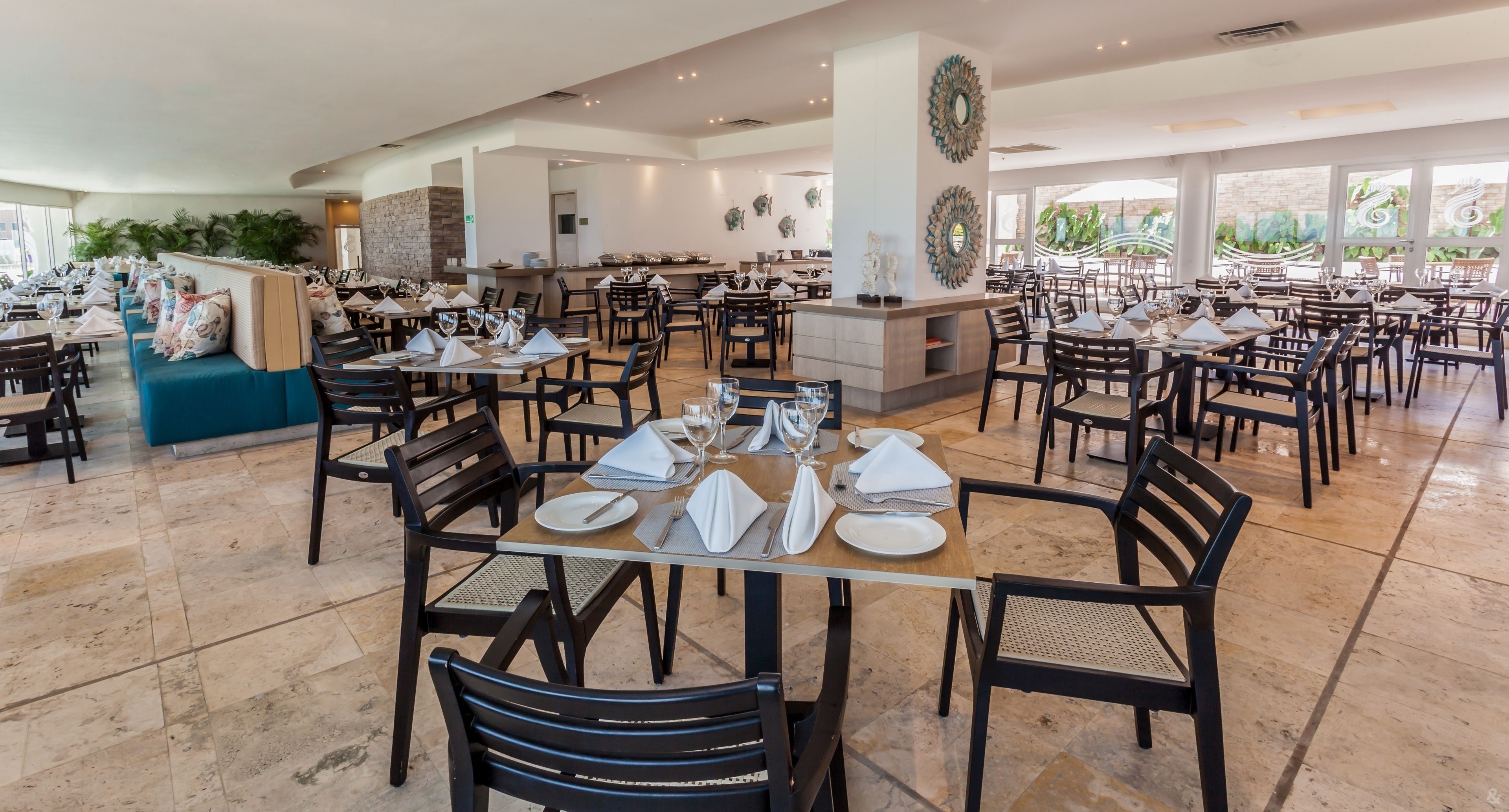 Un restaurante amplio con múltiples mesas y sillas, una zona de bar central y grandes ventanales con vistas a zonas verdes.