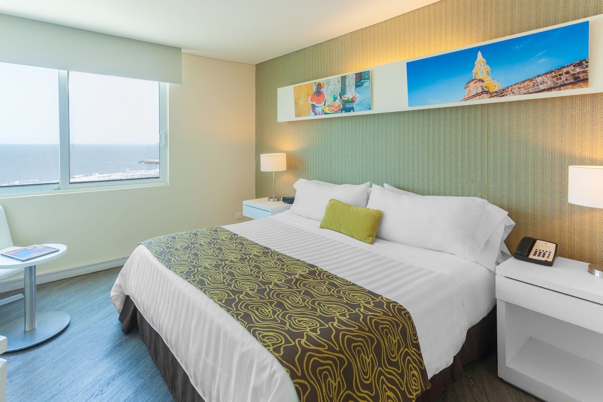 Habitación de hotel con cama, escritorio, silla, ventana con vista al océano y obras de arte en la pared.