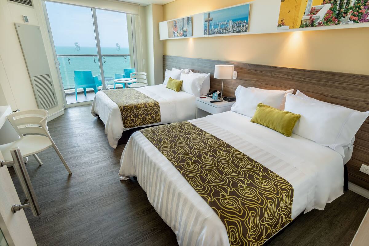 Habitación de hotel con dos camas, balcón con sillas y vista al mar.