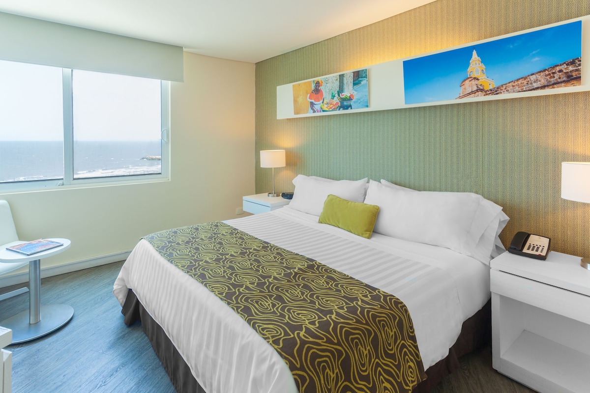 Habitación de hotel con cama, escritorio, silla, ventana con vistas y obras de arte en la pared.