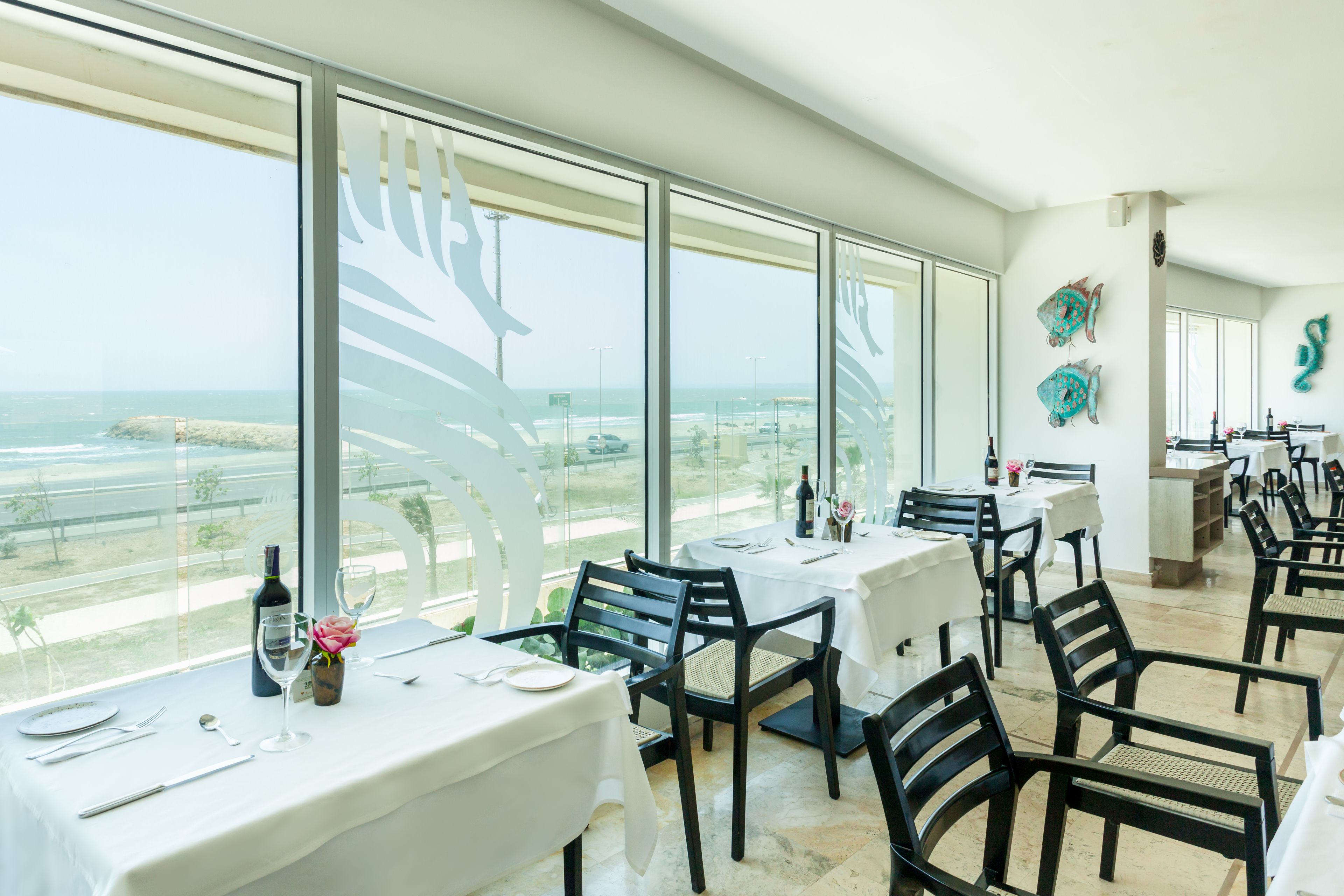 A dining area with tables set for two, large windows with a view of the ocean, and decorative fish wall art.