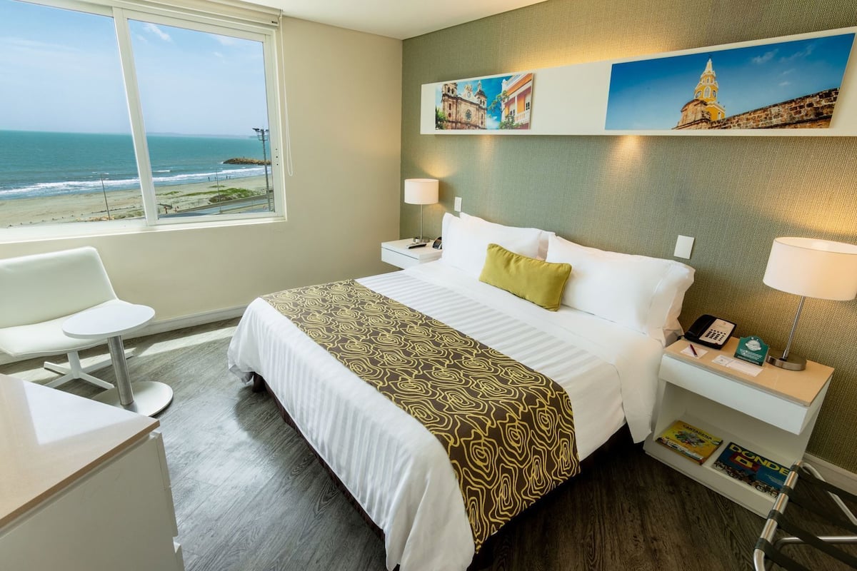Habitación de hotel con cama, escritorio, silla, mesita de noche y vista al océano.