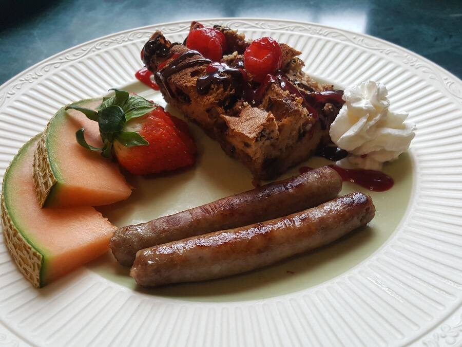 A plate with grilled meat, sausages, a slice of melon, strawberries, whipped cream, and a chocolate drizzle.