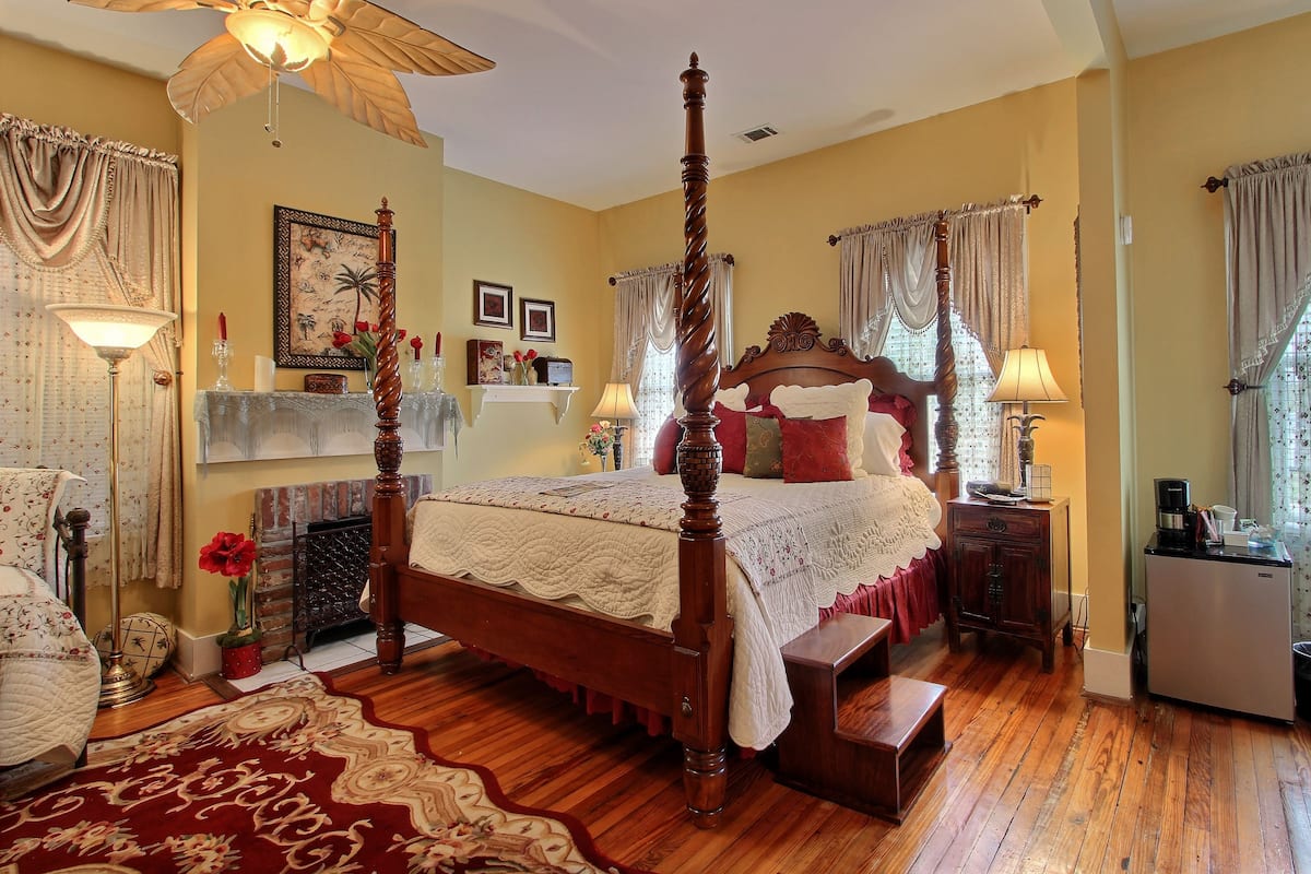 A bedroom with a four-poster bed, a wooden floor, a chandelier, and a small desk with a lamp.