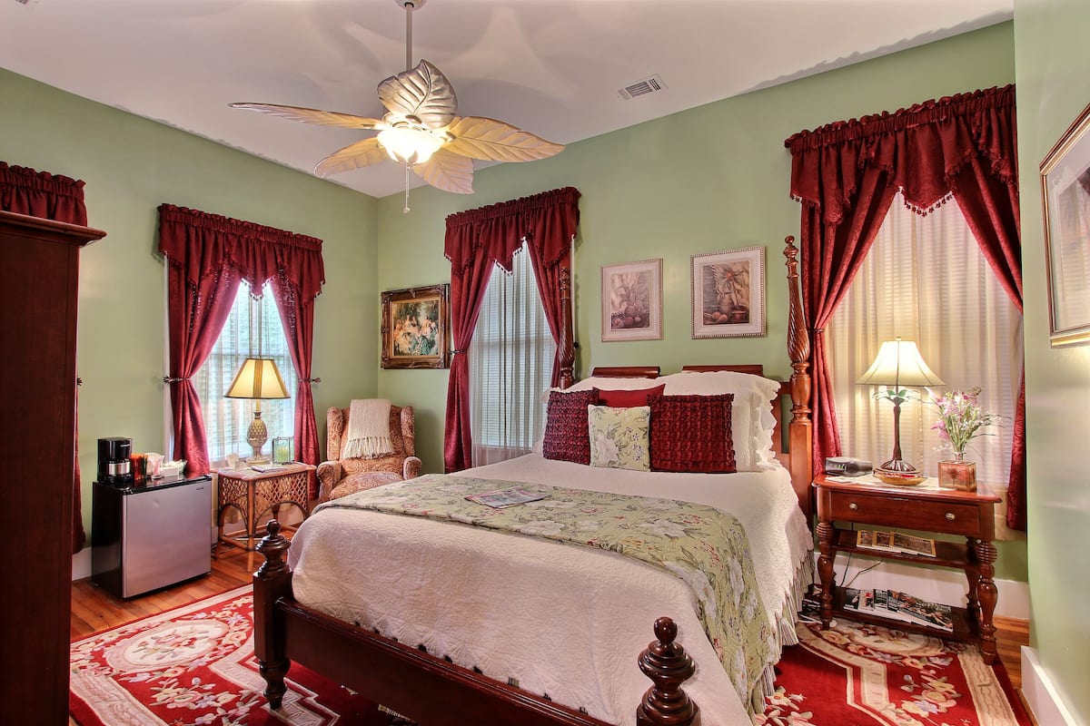A well-appointed bedroom with a large bed, a desk with a lamp, and a small refrigerator.