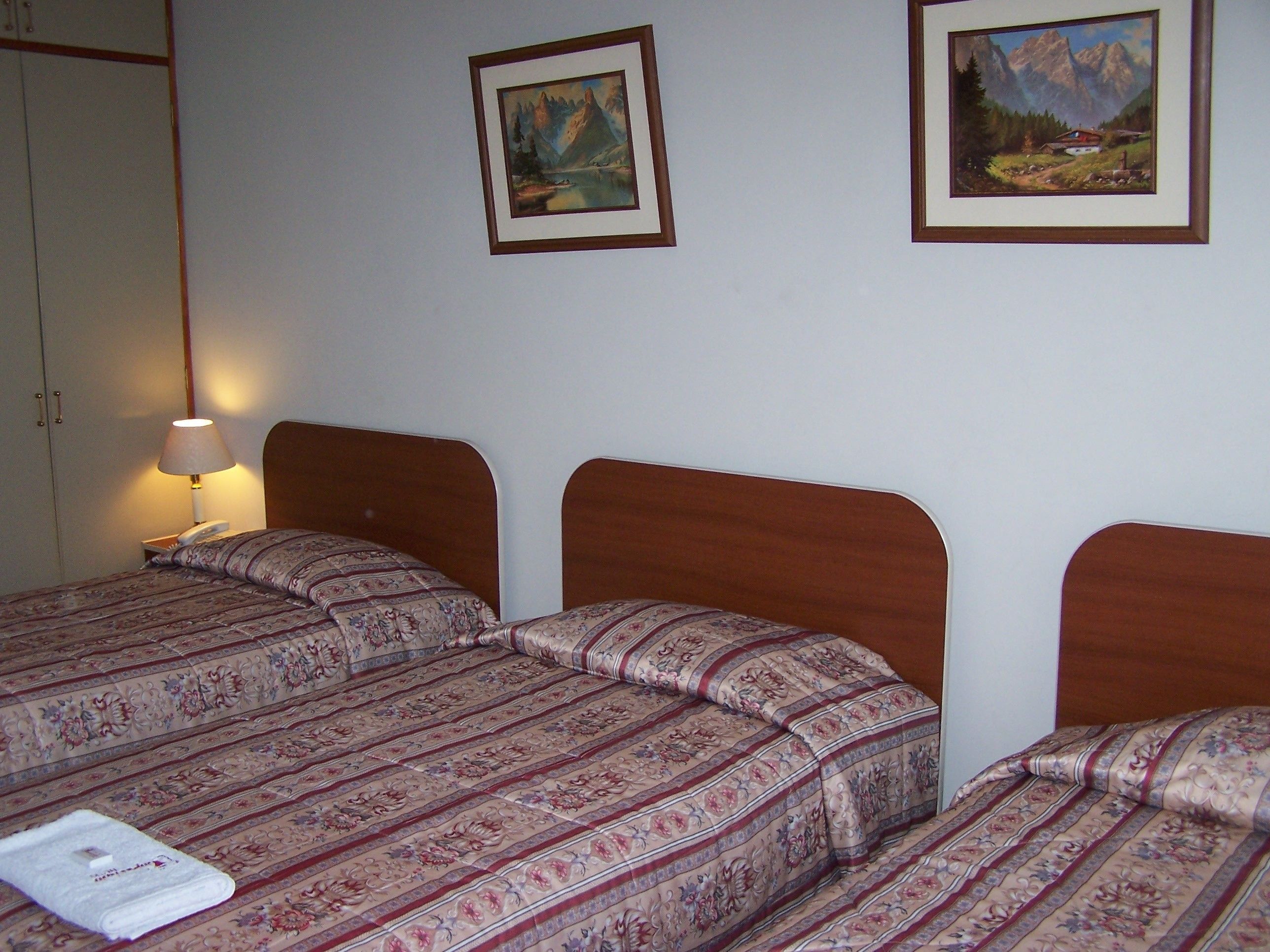 A hotel room with two beds, a bedside table with a lamp, and two framed landscape paintings on the wall.