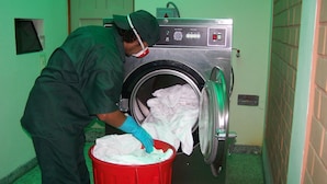 A person in a green uniform loading white linens into a red laundry bin next to a front-loading washing machine in a room with green walls. - Imperium Hotel Lima (Lima)