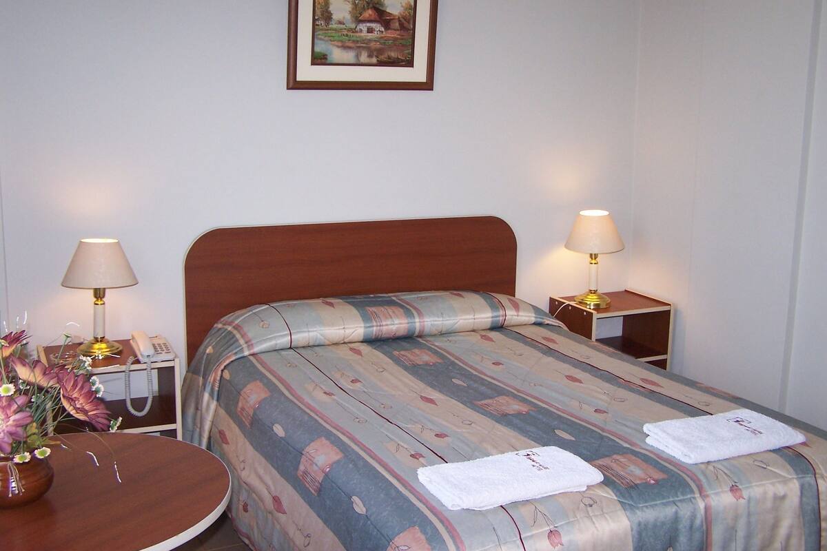 A hotel room with a wooden headboard, a bed with striped bedding, two bedside tables with lamps, a vase with flowers, and a framed picture on the wall.
