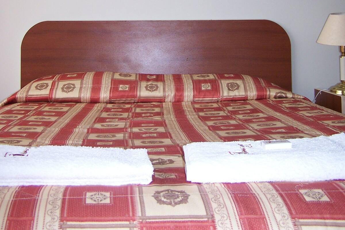 A neatly made bed with a patterned bedspread and two towels, a framed landscape painting on the wall, and a bedside lamp.