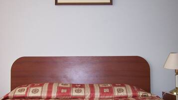 A neatly made bed with a patterned bedspread and two towels, a framed landscape painting on the wall, and a bedside lamp.