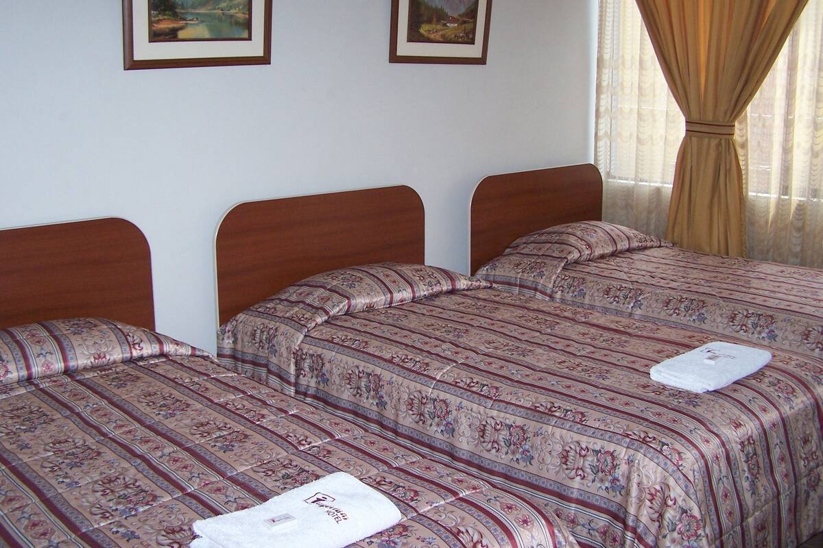 A hotel room with three wooden beds, each with floral-patterned bedding. There are two framed landscape paintings on the wall.
