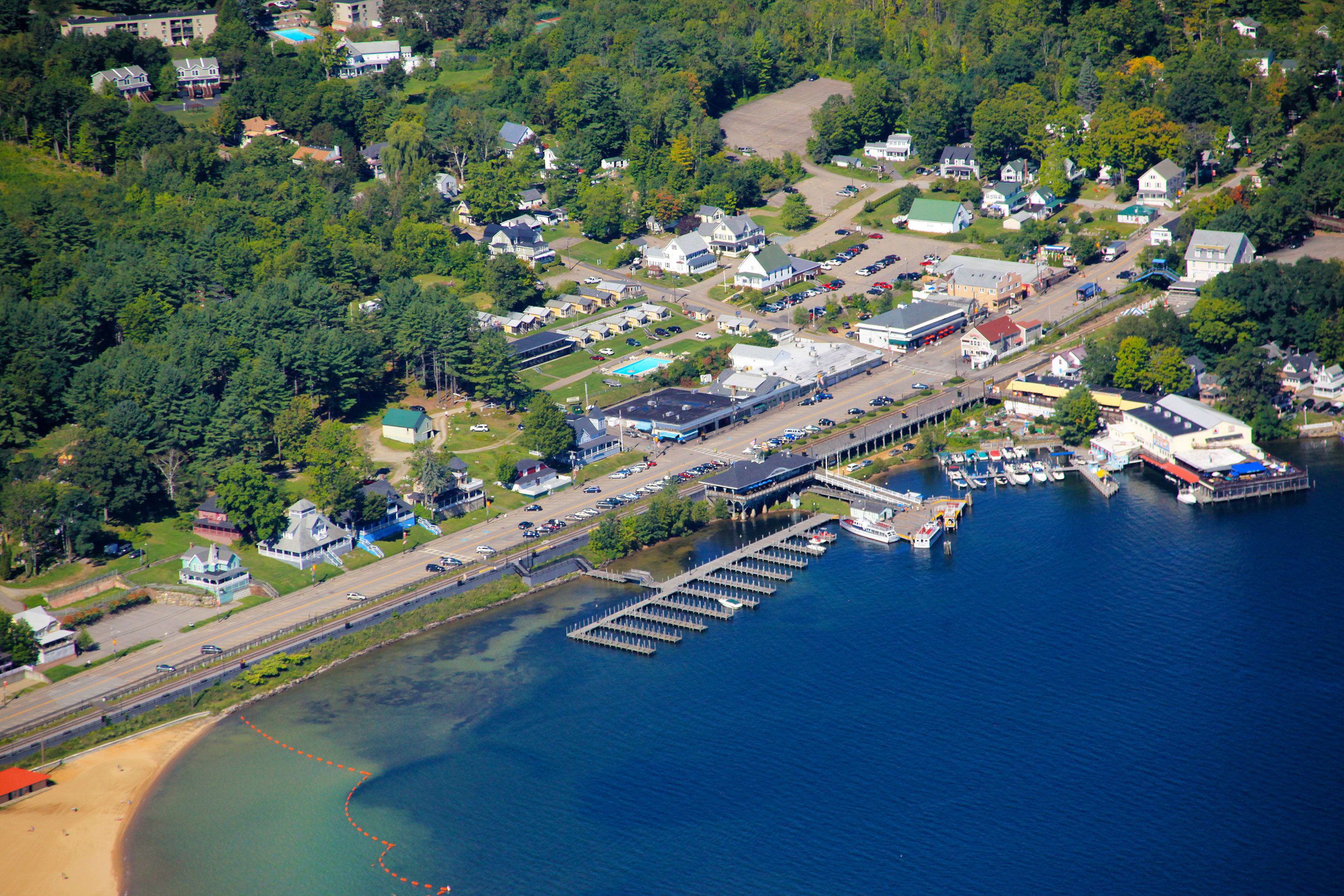Vue aérienne d’une ville située au bord d’un lac, avec un port de plaisance, des immeubles résidentiels et une route menant au rivage.