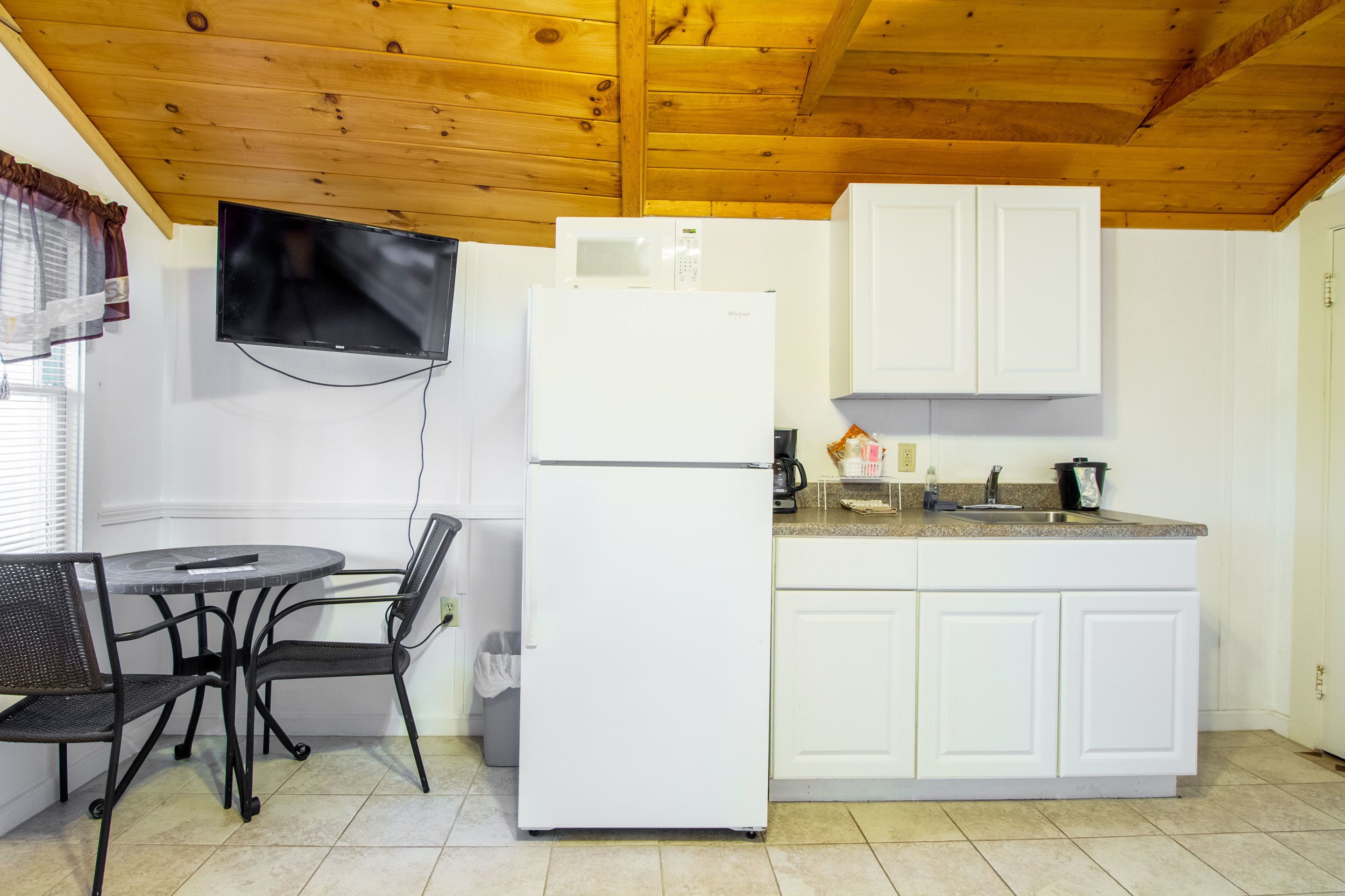 A compact kitchen with a refrigerator, microwave, and white cabinets. There is a small dining table with chairs and a TV mounted on the wall.