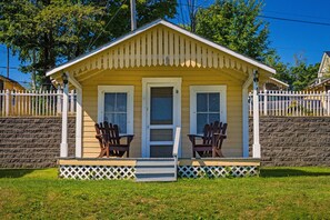 Uma casa pequena, térrea, com varanda, duas cadeiras Adirondack e cerca branca de piquete.