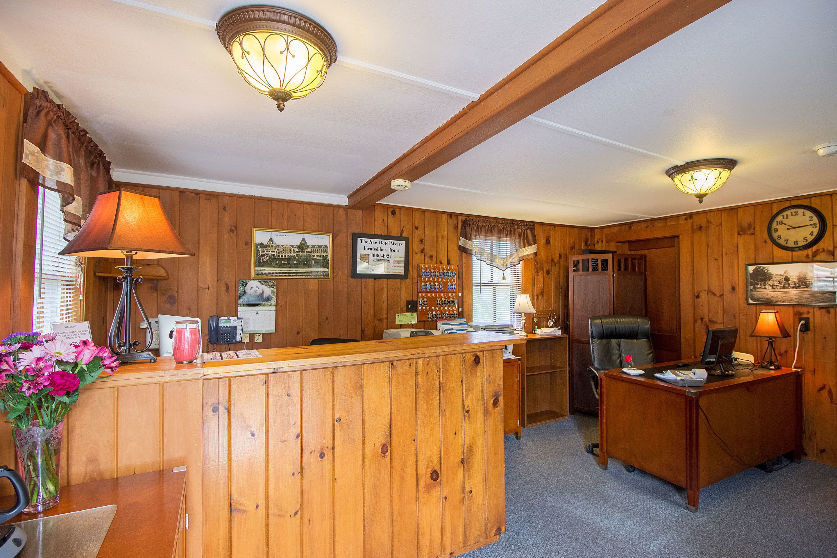 Un comptoir d’accueil en bois, avec un téléphone, un ordinateur et une lampe. Des tableaux encadrés ornent le mur, et il y a aussi une horloge.