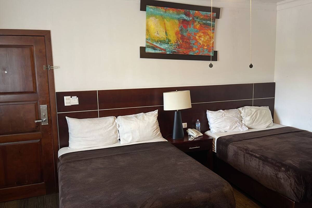 A hotel room with two beds, a nightstand with a lamp, a painting on the wall, and an air conditioning unit.