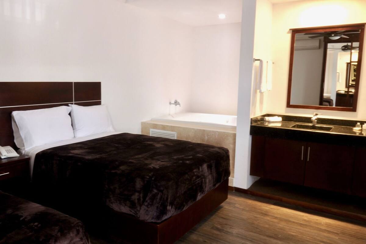 A hotel room with a bed, bedside table, lamp, and a bathroom with a sink and mirror.