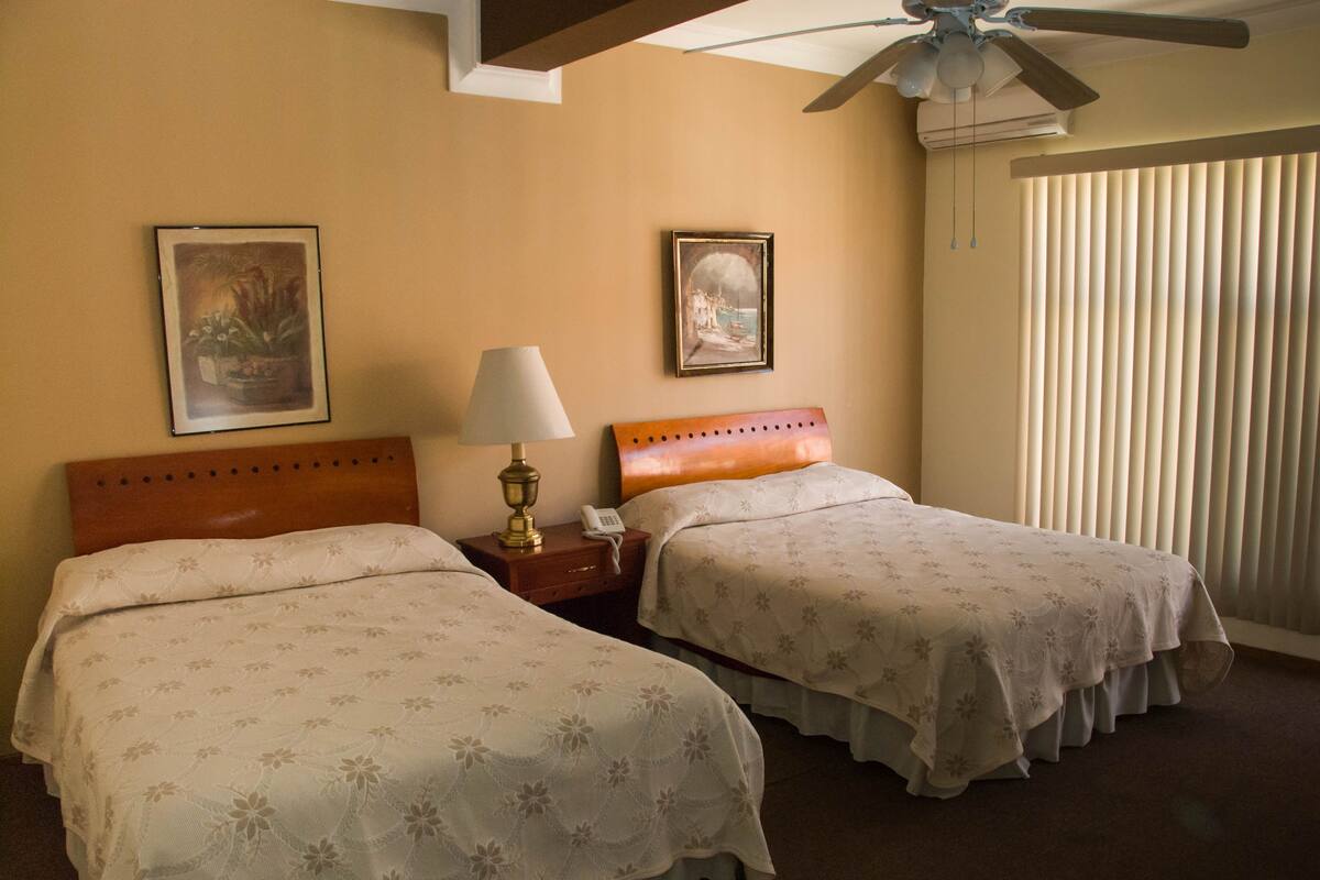A hotel room with two beds, a nightstand with a lamp, a ceiling fan, and a window with blinds.