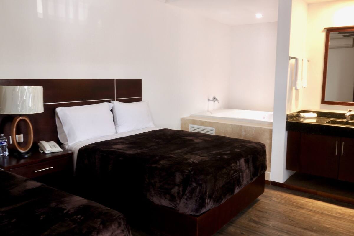 A hotel room with a bed, a bedside table with a lamp, a phone, and a water bottle. There is a bathroom with a sink and a mirror.