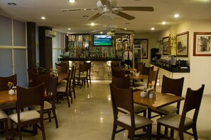 Restaurante com várias mesas e cadeiras, área de bar com televisão e ventilador de teto.