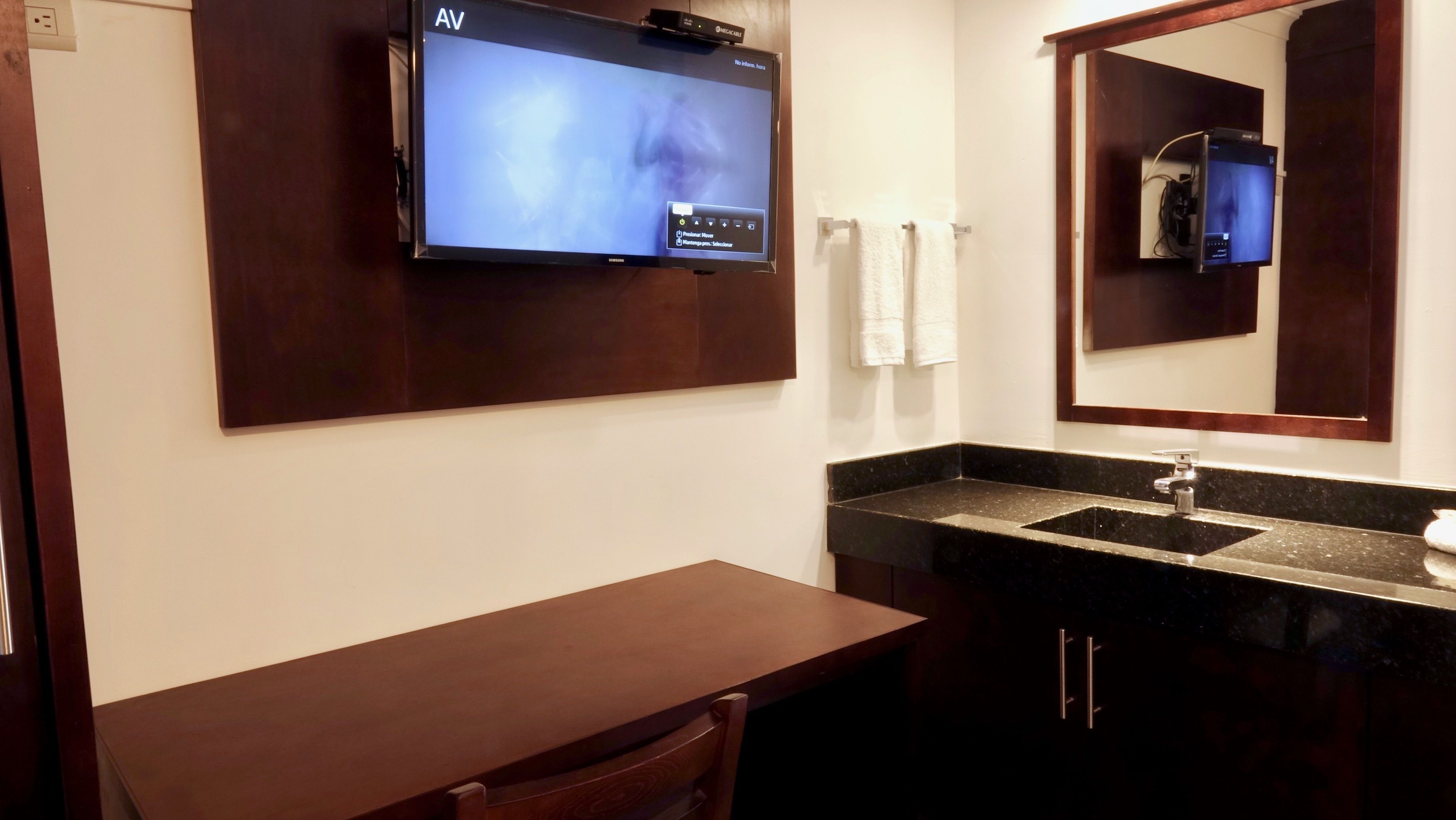 A bathroom with a flat-screen TV mounted on the wall, a wooden bench, a sink with a dark countertop, and a mirror above the sink.