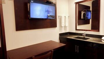 A bathroom with a flat-screen TV mounted on the wall, a wooden bench, a sink with a dark countertop, and a mirror above the sink.
