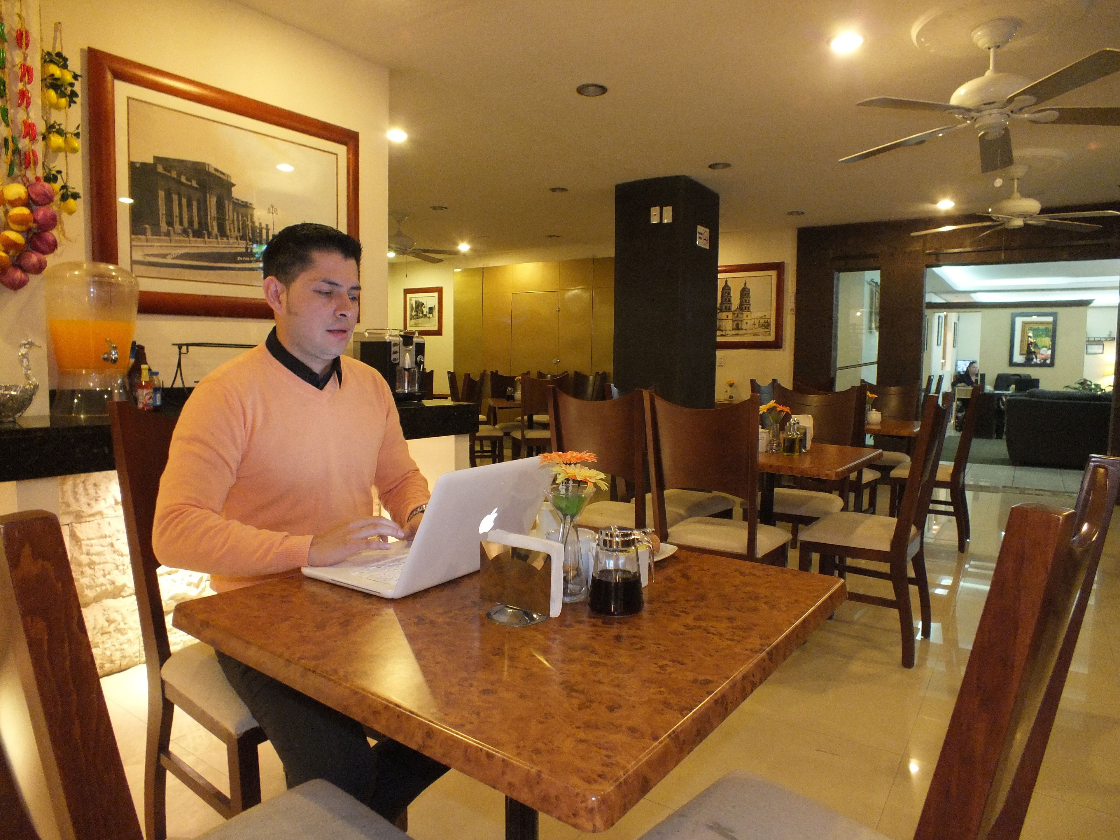 Um homem trabalha num computador portátil numa mesa de madeira numa sala de jantar com cadeiras e uma máquina de café.