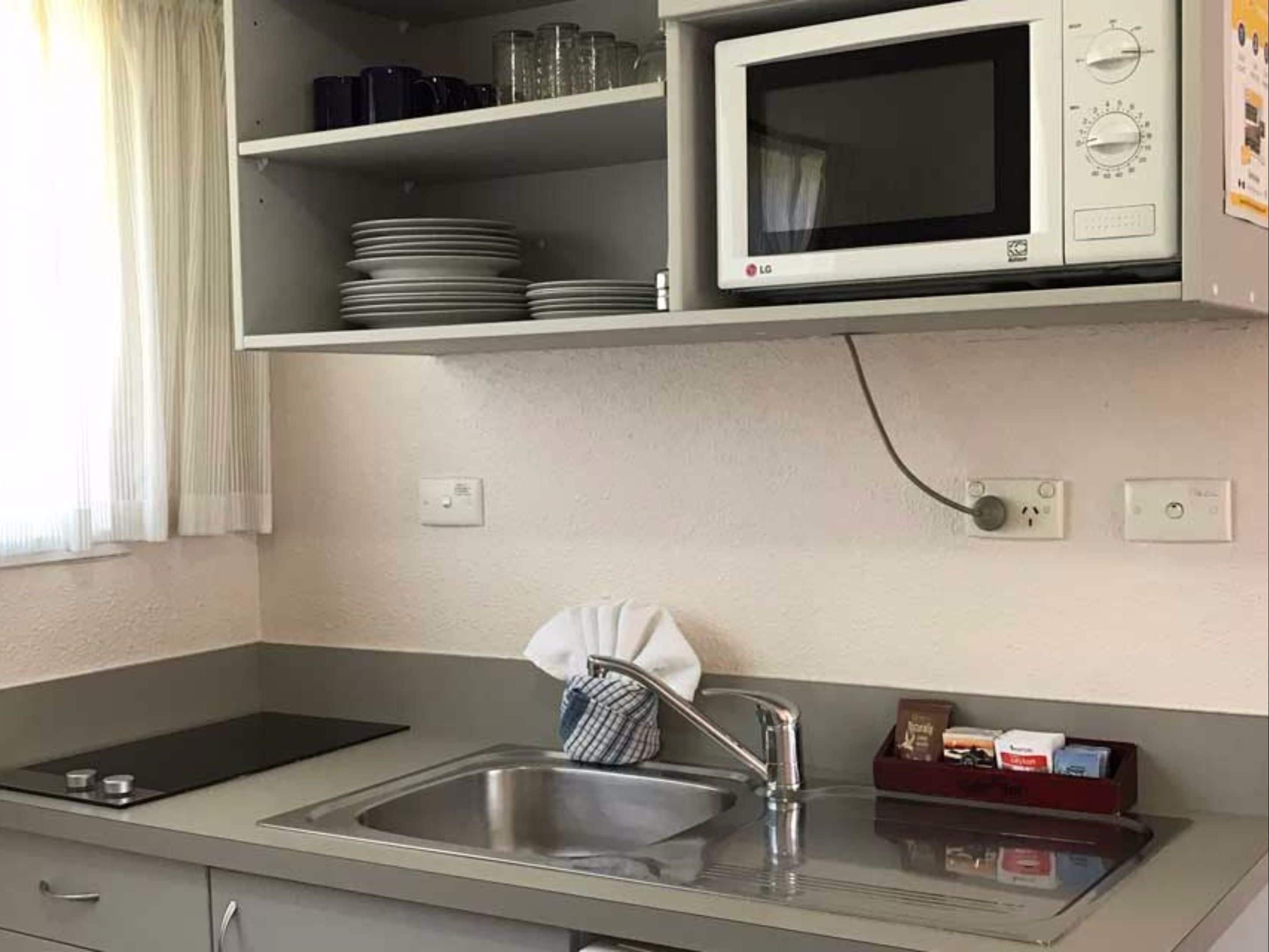 A kitchen with a microwave, sink, and a shelf above the counter holding plates and cups.