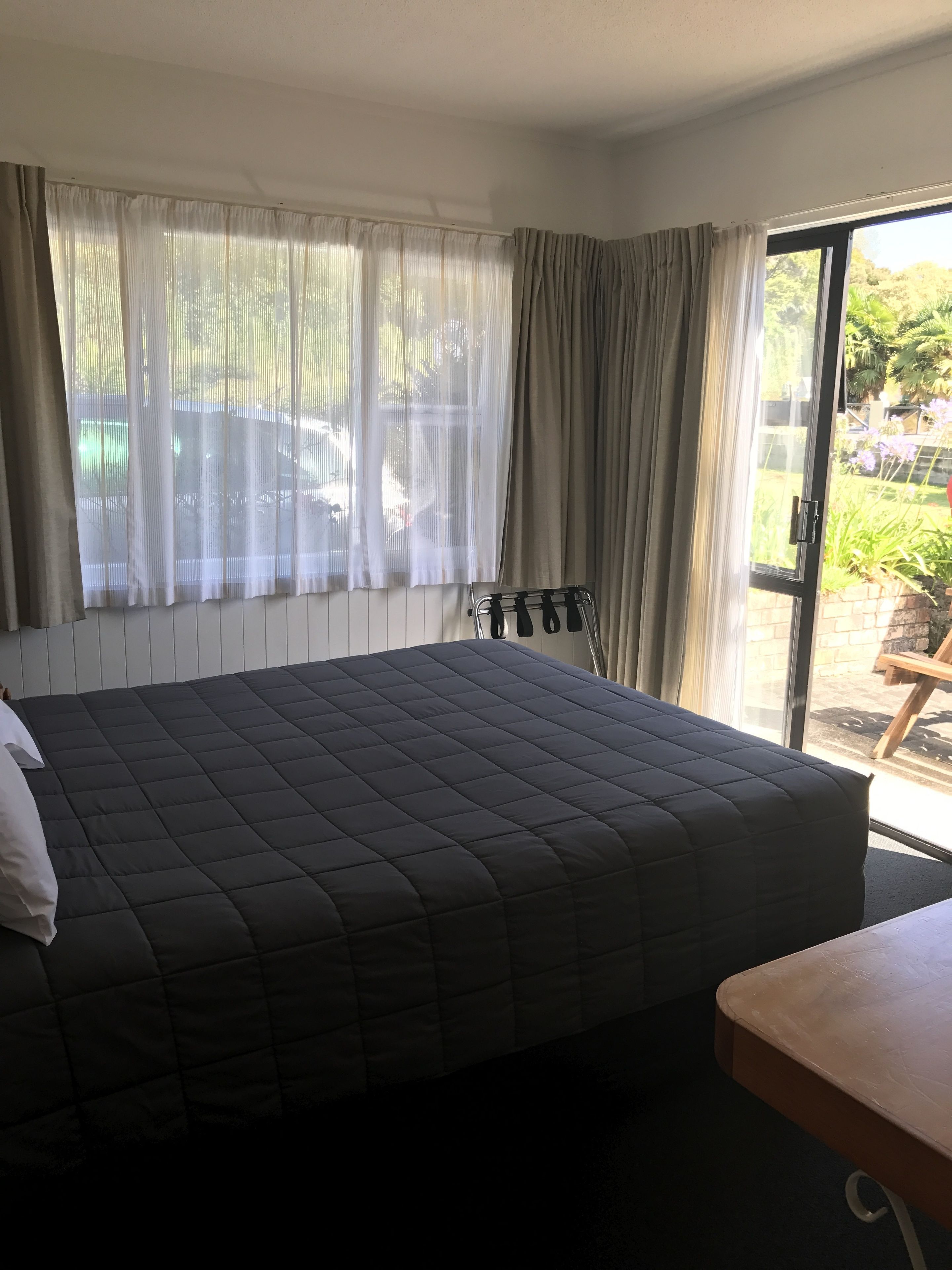 A hotel room with a large bed, a wooden table, and a view of the outdoors through a window with curtains.