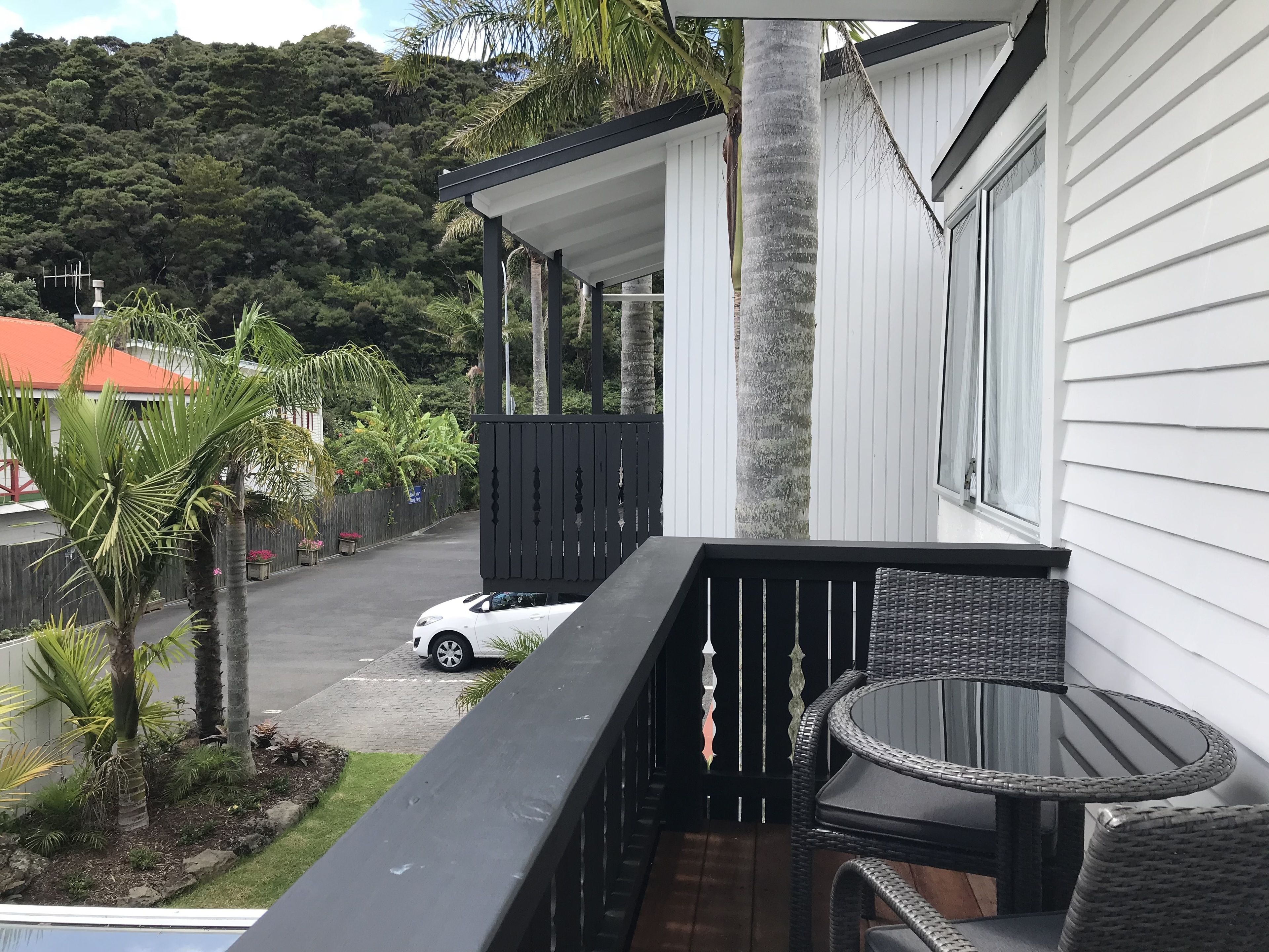 A balcony with a wicker table and chairs, a white car parked outside, and a view of a lush green hill in the background.