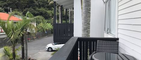A balcony with a wicker table and chairs, a white car parked outside, and a view of a lush green hill in the background.