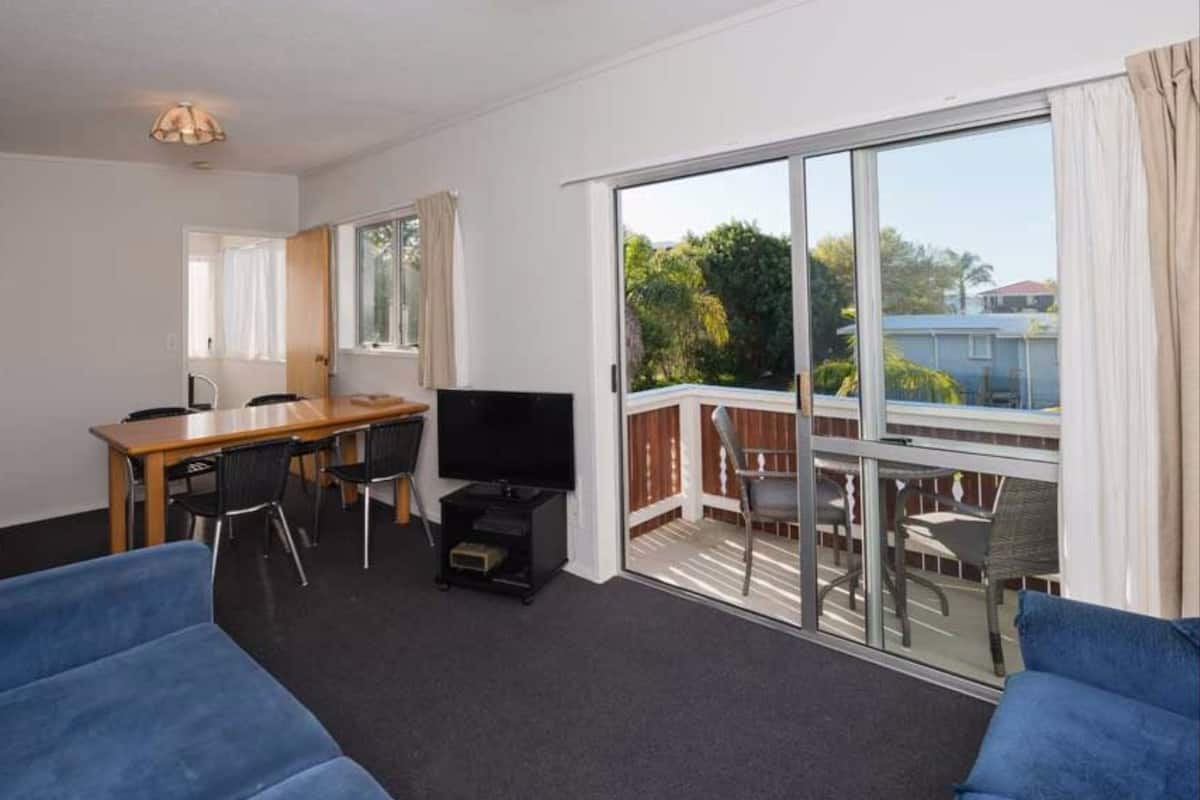 A room with a TV, a wooden table, and chairs, a balcony with a view of greenery, and a large window.