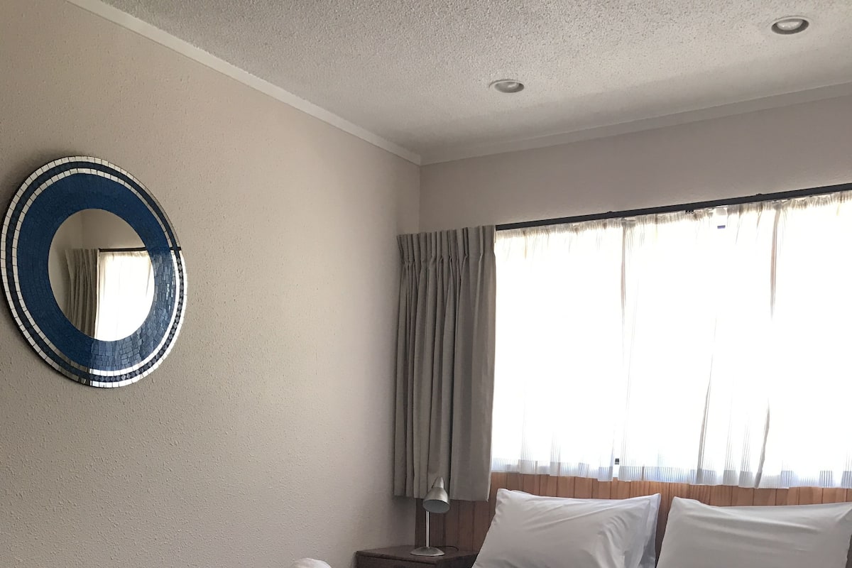 A hotel room with a large bed, a round mirror, a nightstand, and a window with curtains.
