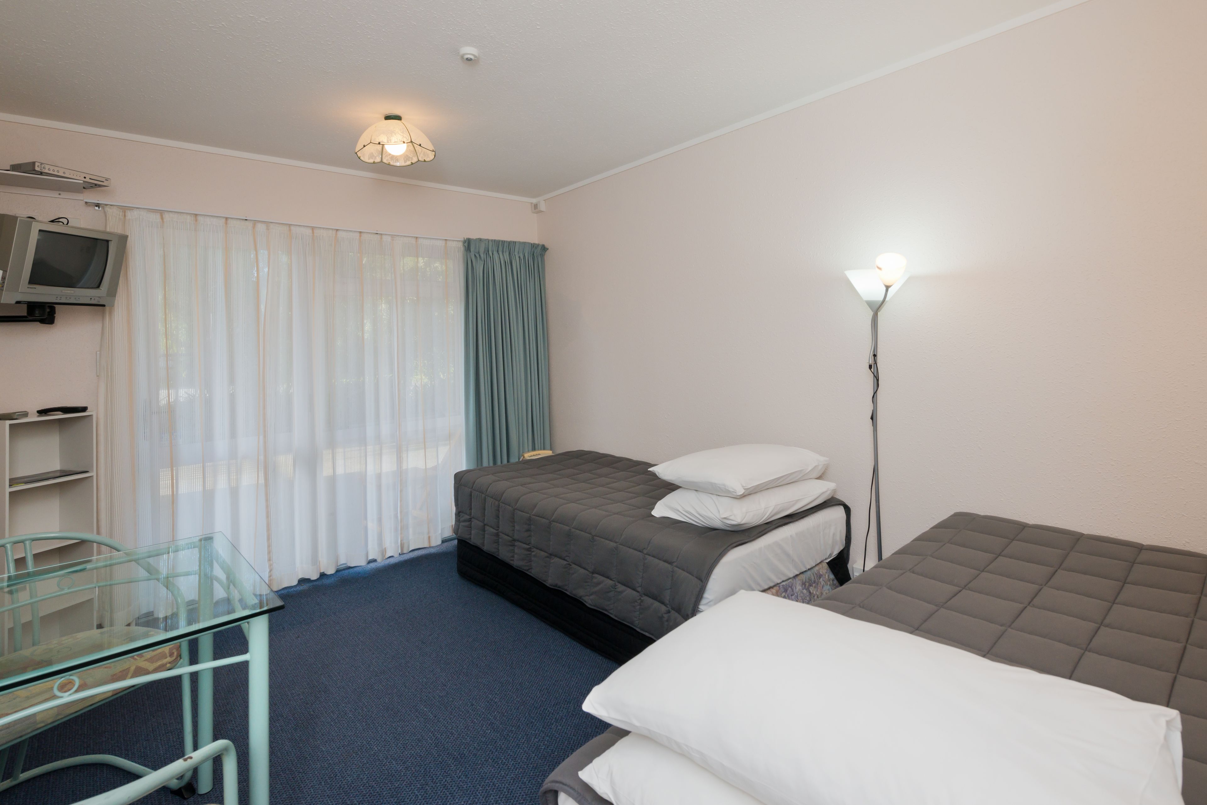 Habitación de hotel con una cama individual, una mesita de noche, un escritorio pequeño con una silla, un televisor y una lámpara de pie.