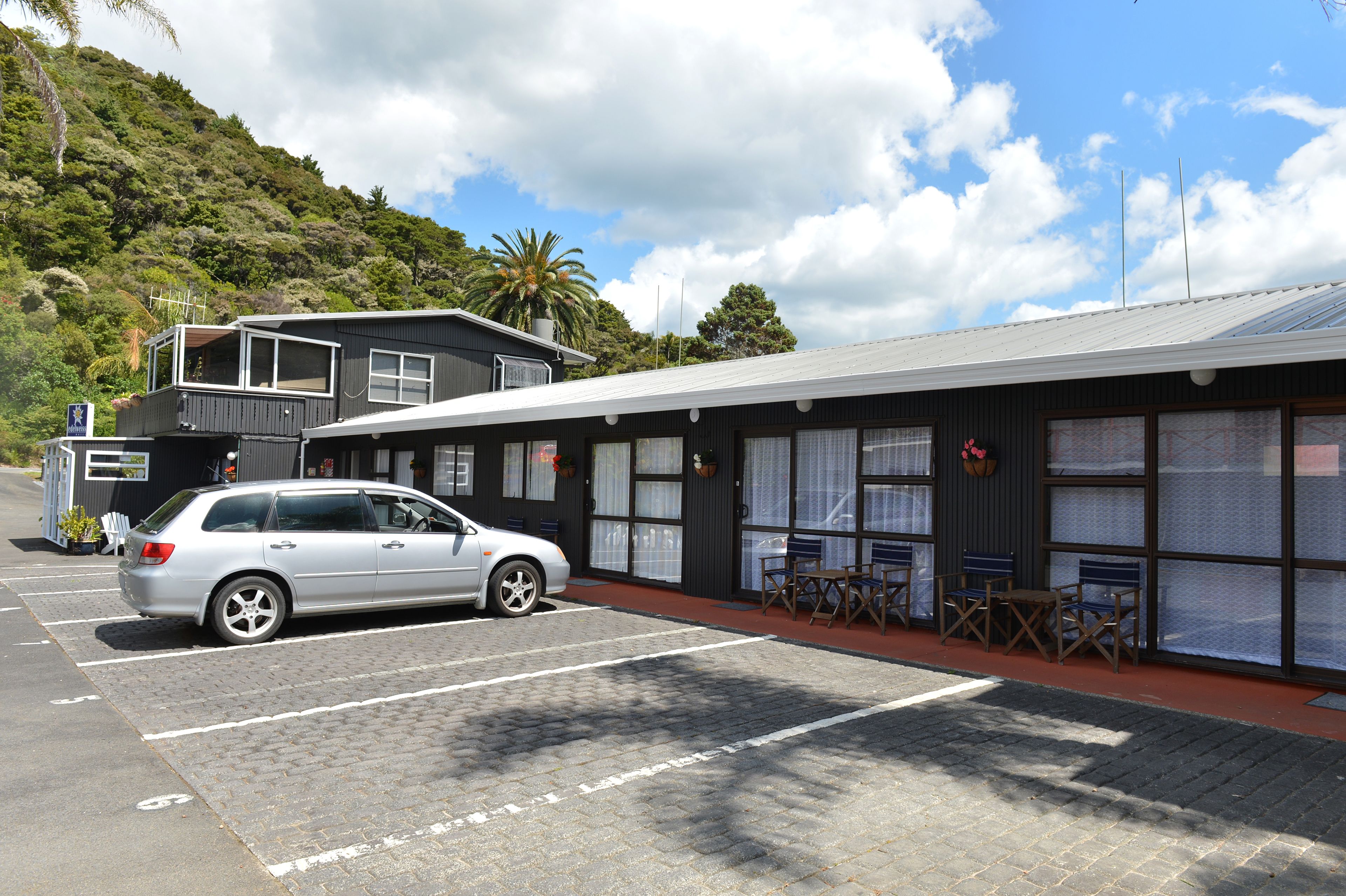 A parking lot with a silver car, a building with large windows and a flat roof, and a hillside in the background.