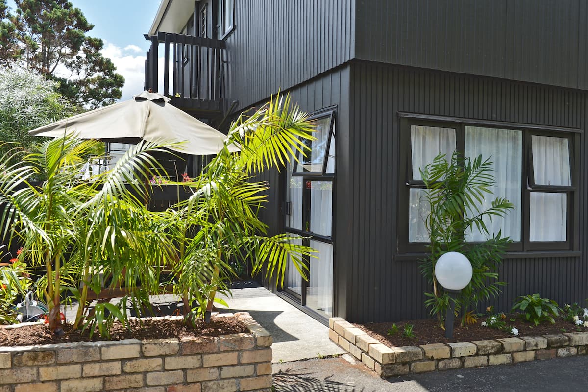 A modern building with a dark exterior, large windows, and a well-maintained garden with palm trees and a patio umbrella.