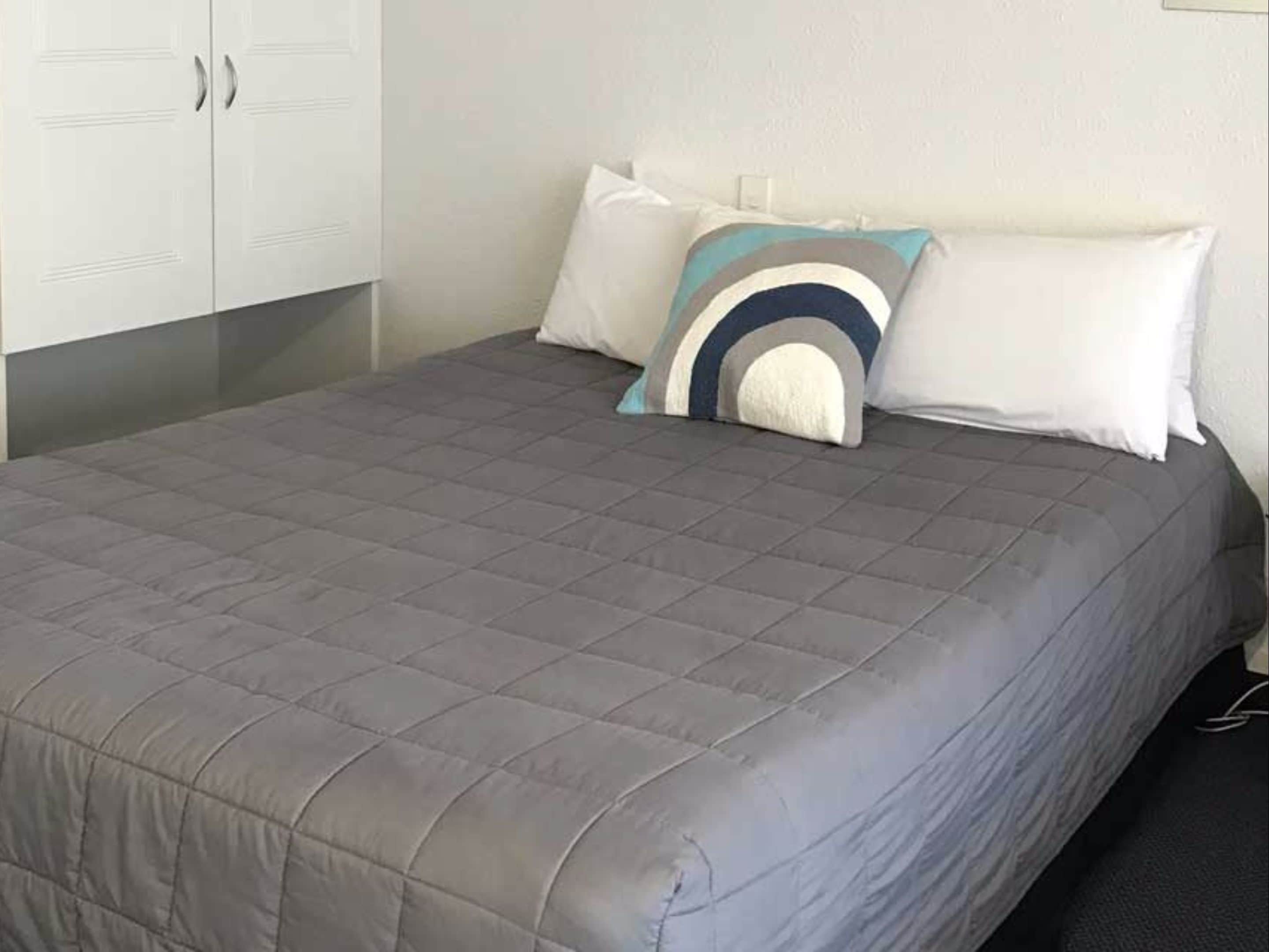 Una cama bien hecha con un edredón gris, almohadas blancas y una almohada decorativa con estampado azul y blanco.