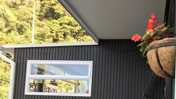 A dark wooden wall with a window, two white chairs, and a hanging basket with red flowers. - Edelweiss Motel (Paihia)