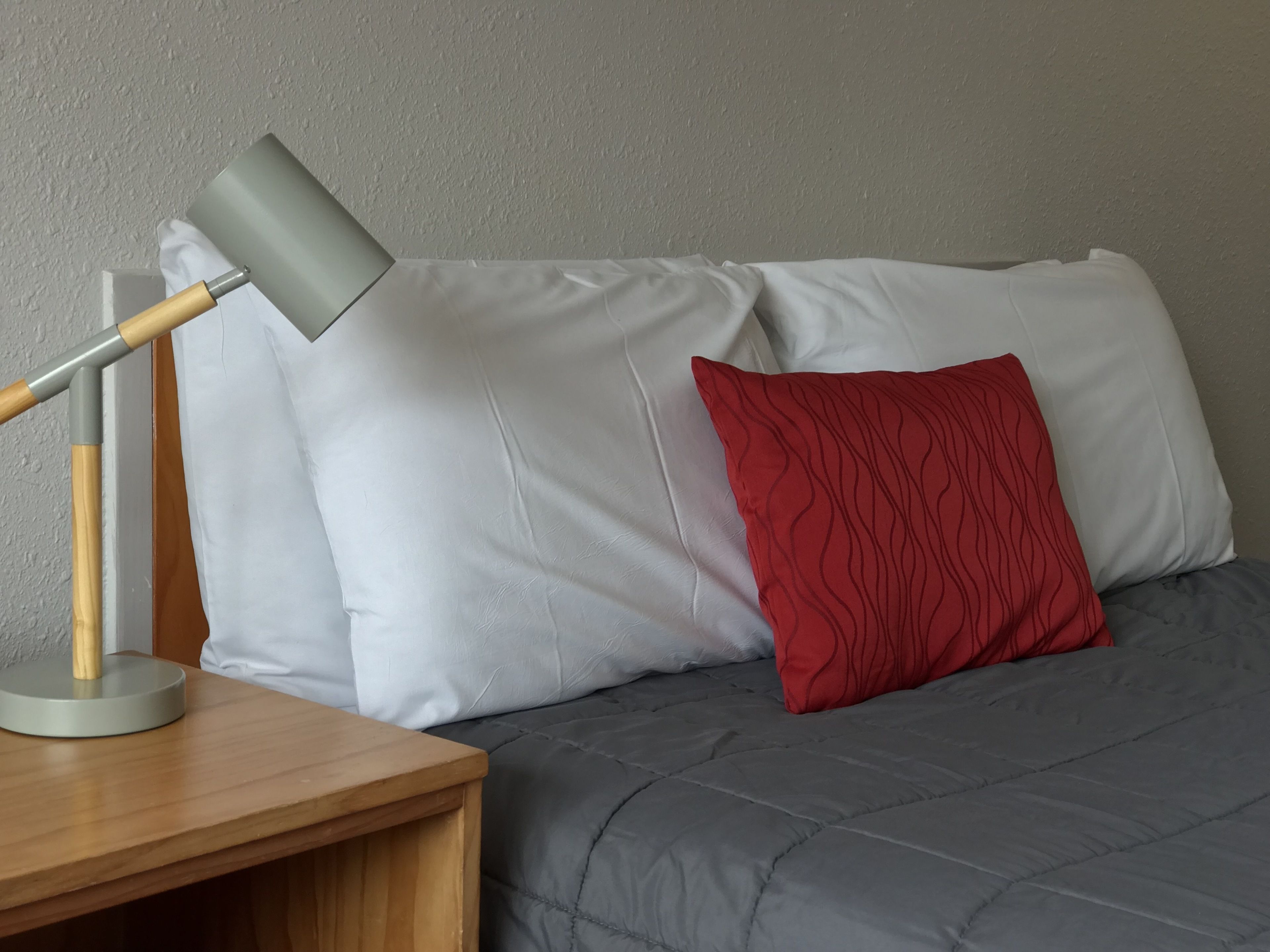 A bed with a grey duvet and a red pillow, a wooden bedside table with a lamp, and a plain wall in the background.